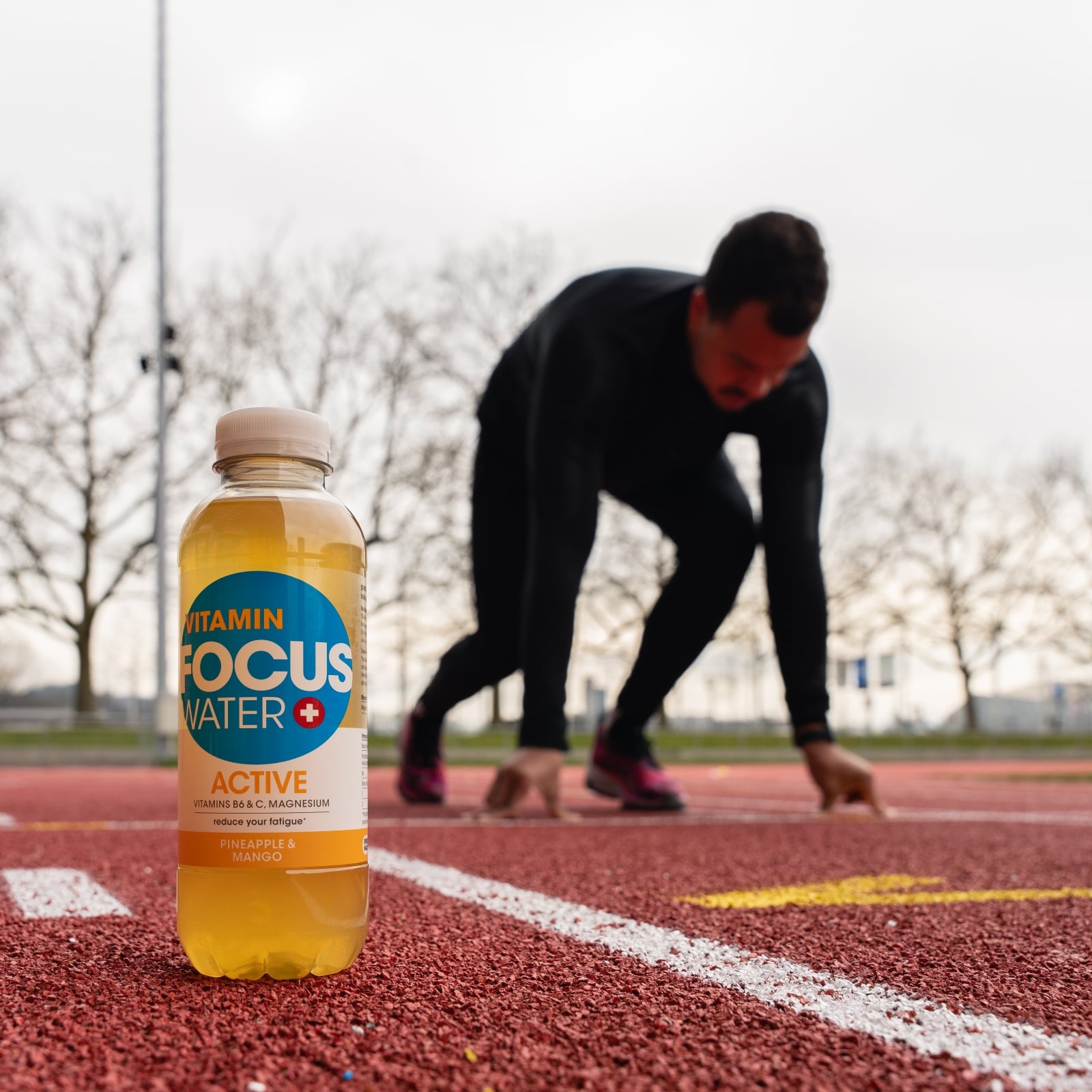 Eine Focuswater Active Vitaminwasser Flasche steht auf dem Boden vor einem Sprinter der sich in Position bringt