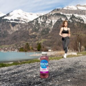 Eine Flasche Focuswater Care Vitaminwasser steht auf einem Weg und im Hintergrund läuft eine junge Frau