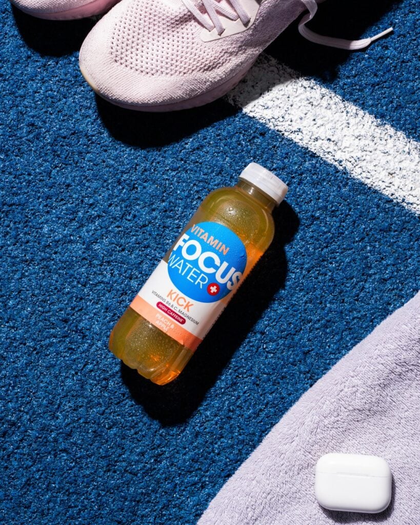 Focuswater Kick Flasche auf blauem Untergrund neben Sportschuhen.