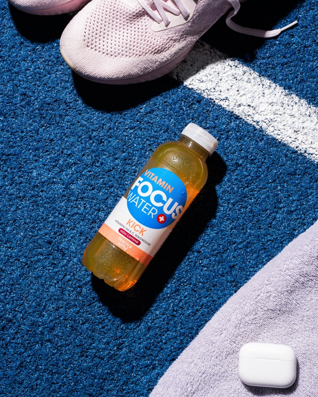 Focuswater Kick Flasche auf blauem Untergrund neben Sportschuhen.