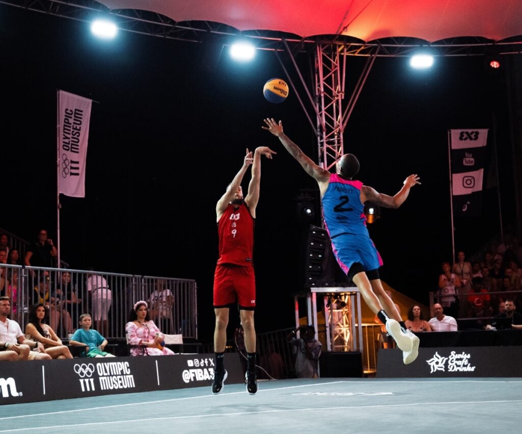 Basketballspiel bei Nacht, zwei Spieler in roter und blauer Kleidung.