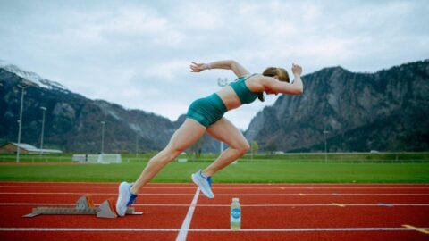 Eine Athleting beginnt ihren Sprint auf einer roten Laufbahn und im Hintergrund sind Berge zu sehen