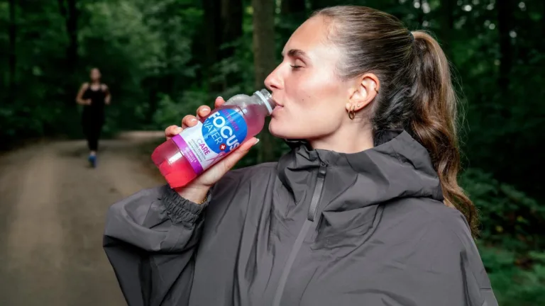 Frau in grauer Jacke trinkt Focuswater Care im Wald.