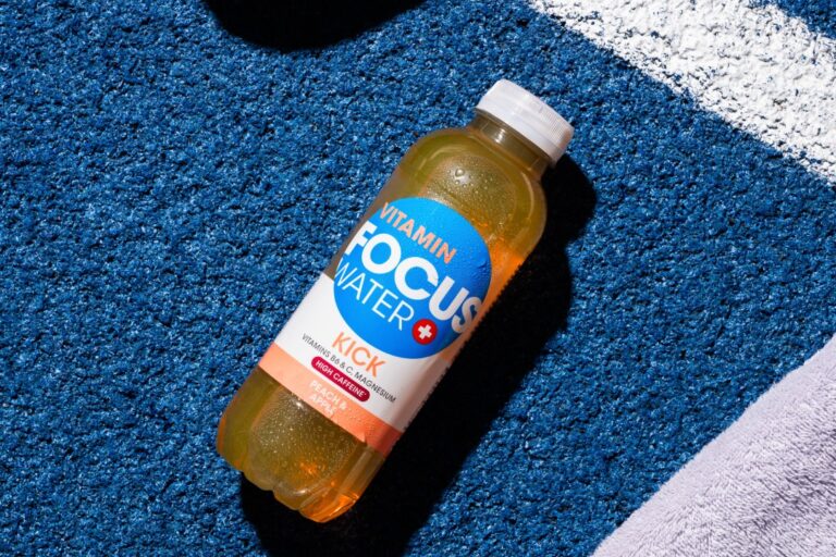 Focuswater Kick Flasche auf blauem Untergrund neben Sportschuhen.